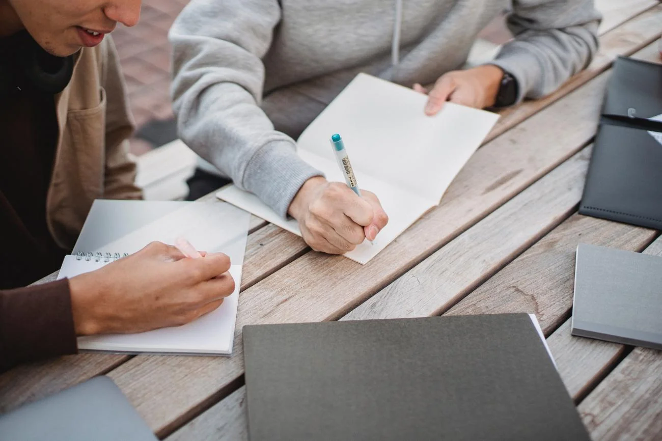 Two students journaling assignment notes at a wooden desk
